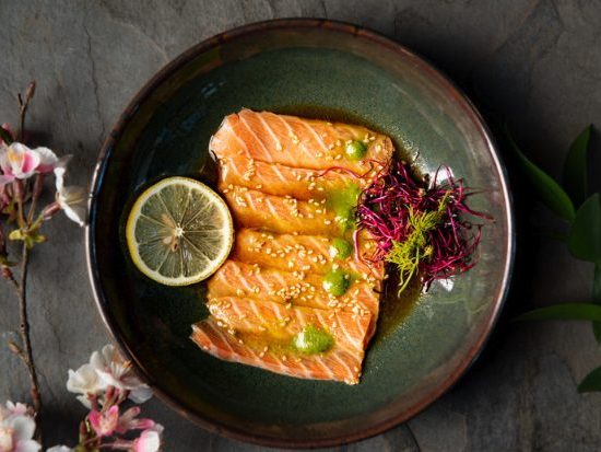 Pieces of salmon on a dark place with a dollop of wasabi on them. A lemon slice and cherry blossoms are also in the photo.