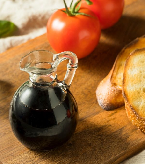 Balsamic vinaigrette in a small glass vase on a wooden cutting board along with toasted golden bread, tomatoes and basil.