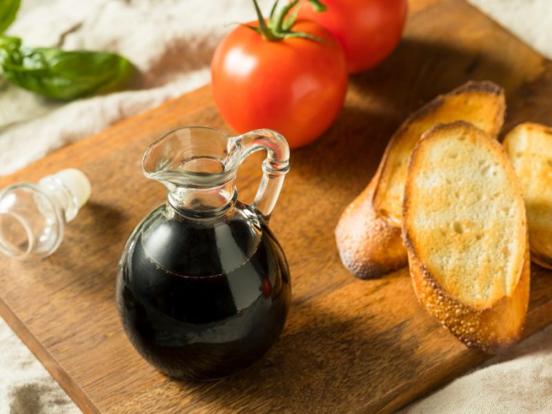 Balsamic vinaigrette in a small glass vase on a wooden cutting board along with toasted golden bread, tomatoes and basil.
