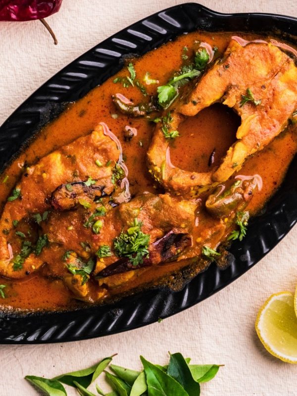Fish covered in simmering red sauce in a black pan surrounded by lemon parsley, onions, peppers, and spices.